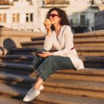 Beautiful stylish smiling lady in sunglasses sitting on city stairs with cellphone and apple